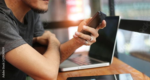 Man in casual wear with mobile smartphone in hand and laptop on table, working in cafe
