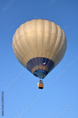 Ogromny szary z niebieskimi dodakami balon na ogrzane powietrze unosi się na niebie z doczepionym wiklinowym koszem z załogą, przy balonie biało-czerwona flaga Polski, widok z ziemi