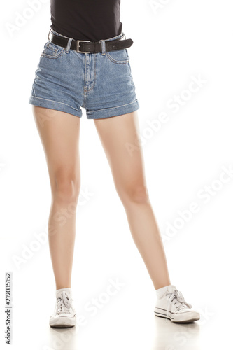 Female legs in short jeans and sneakers standing on white background