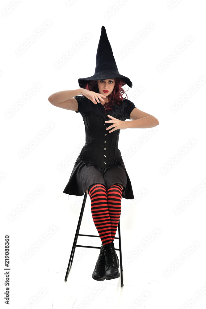 full length portrait of red haired girl wearing long black cloak, pointy hat and witch costume. seated pose on chair, isolated on white studio background. full length portrait of red haired girl wearing long black cloak, pointy hat and witch costume. seated pose on chair, isolated on white studio background.