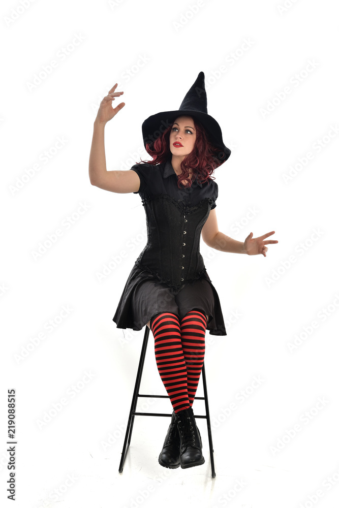 full length portrait of red haired girl wearing long black cloak, pointy hat and witch costume. seated pose on chair, isolated on white studio background. full length portrait of red haired girl wearing long black cloak, pointy hat and witch costume. seated pose on chair, isolated on white studio background.