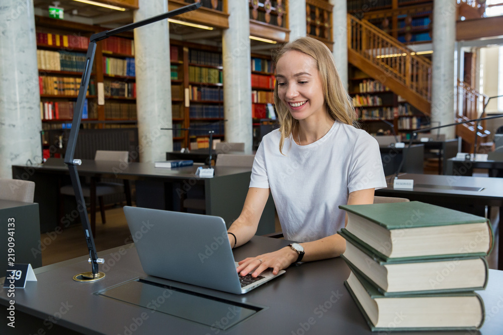 Young girl sitting at a desk in library, working at computer. Smiling student with laptop, studying. Higher education. Young girl sitting at a desk in library, working at computer. Smiling student with laptop, studying. Higher education.