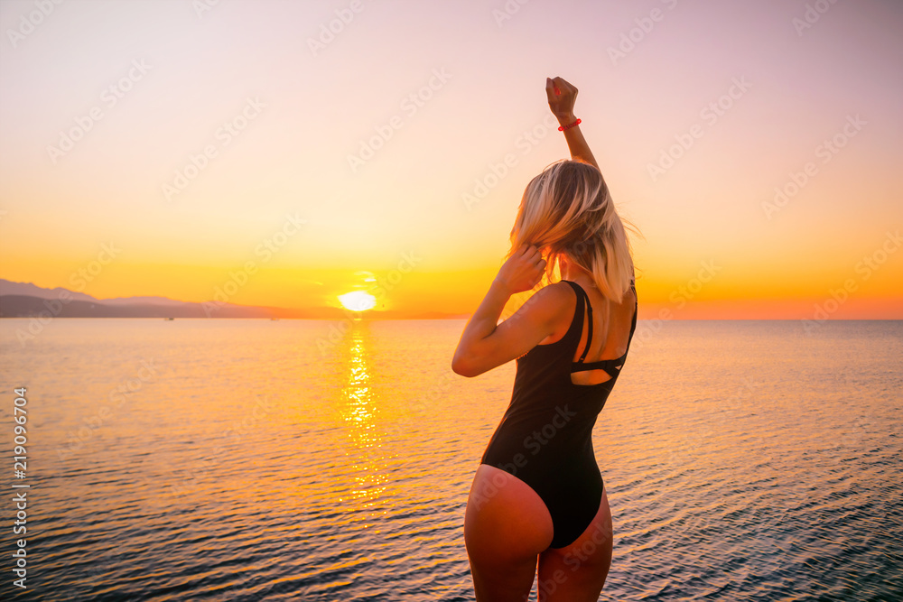 silhouette of a girl with a sexy ass in a black bikini, meets the sunrise on the coast, pulling one hand up, and the second holds the hair from the streams of warm air.
