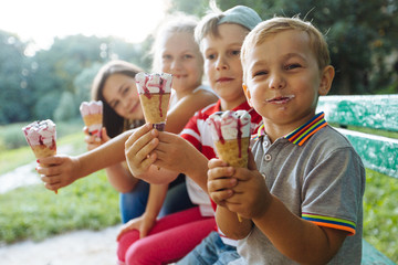 Group of four happy children eating ice cream together outdoor. Photo of happy blond girls with two handsome boys sitting on the bench and smiling at camera. Sun glare effect