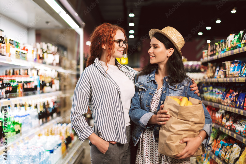 Cheerful girls with paper grocery  bag full of products joyfully looking at each other while spending time in modern supermarket