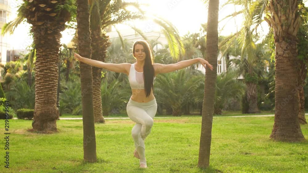 Young athletic woman in white sports outfit performing Yoga Eagle pose outdoor, in park meadow. Young athletic woman in white sports outfit performing Yoga Eagle pose outdoor, in park meadow.