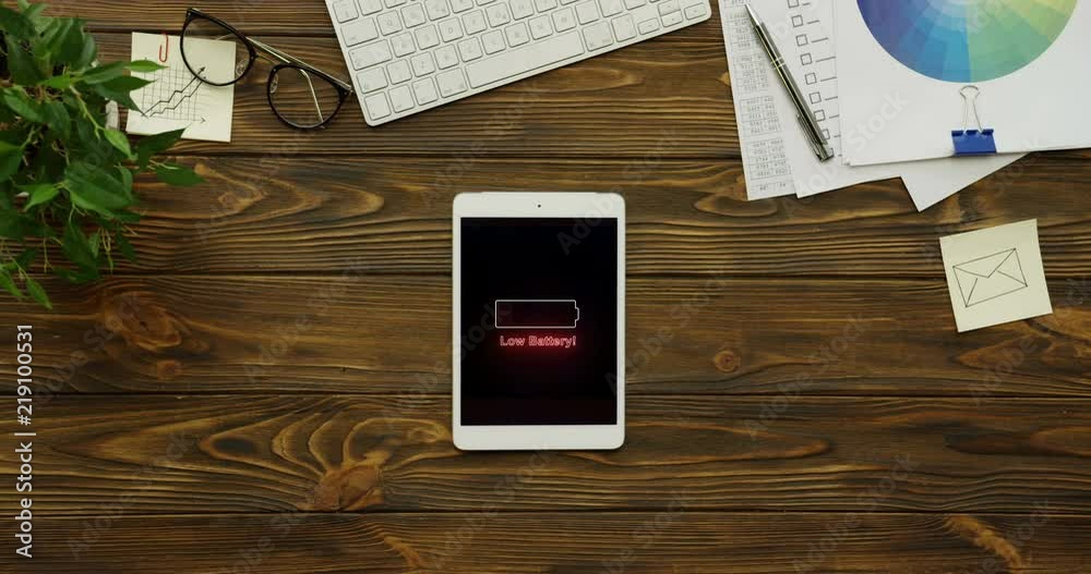 Animated icon of red neon battery blinking as being out of the charge on the vertical black screen of the white tablet device on the wooden desk. Animated icon of red neon battery blinking as being out of the charge on the vertical black screen of the white tablet device on the wooden desk.