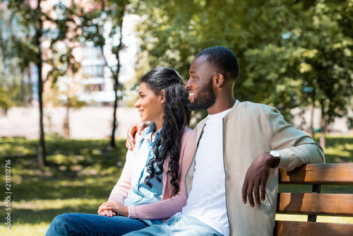 smiling african american couple sitting on wooden bench in park and looking away