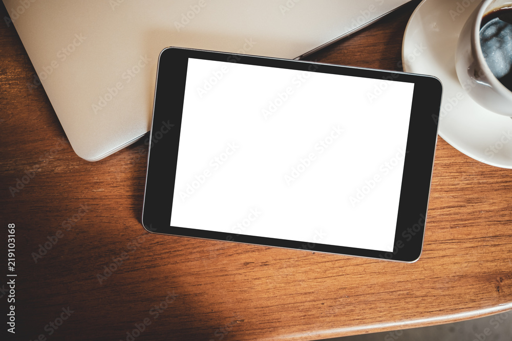 Top view mockup image of a black tablet pc with blank desktop white screen with laptop and coffee cup on wooden table Top view mockup image of a black tablet pc with blank desktop white screen with laptop and coffee cup on wooden table