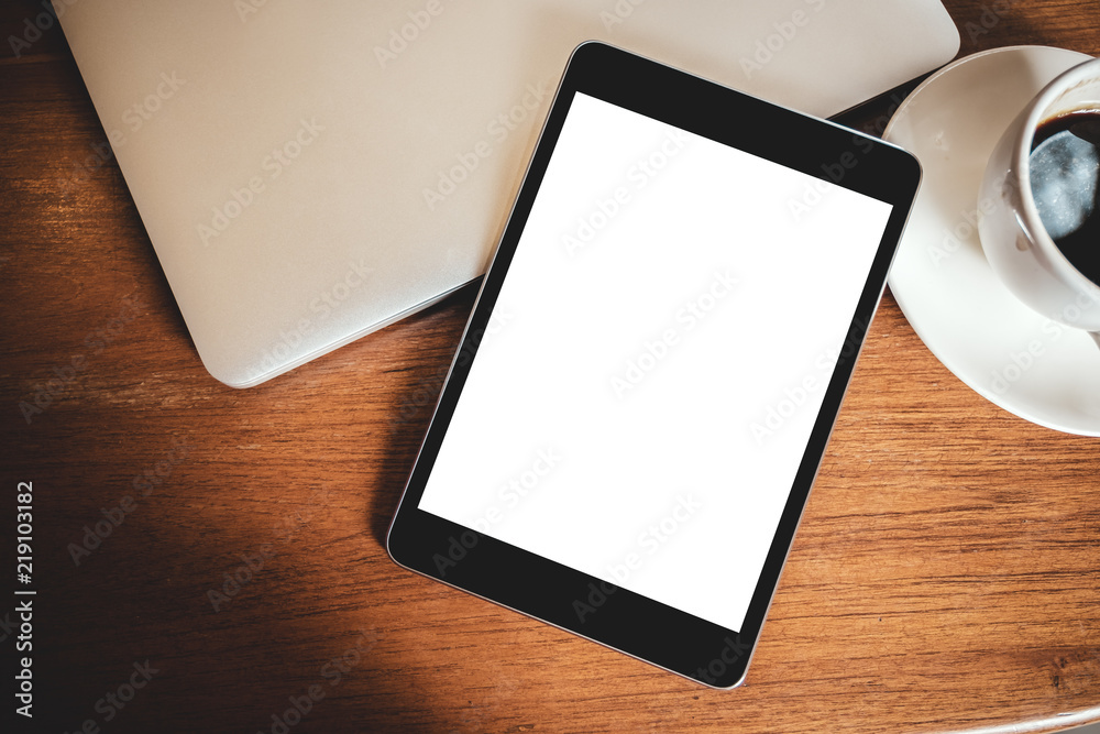 Top view mockup image of a black tablet pc with blank desktop white screen with laptop and coffee cup on wooden table Top view mockup image of a black tablet pc with blank desktop white screen with laptop and coffee cup on wooden table