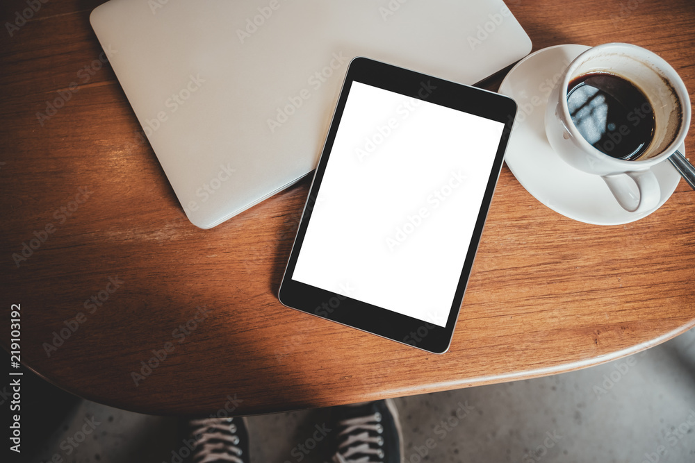 Top view mockup image of a black tablet pc with blank desktop white screen with laptop and coffee cup on wooden table Top view mockup image of a black tablet pc with blank desktop white screen with laptop and coffee cup on wooden table