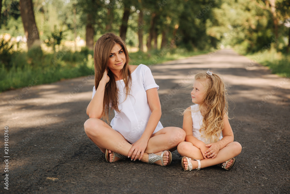 Attractive Mom and blonde hair daughter sits on road near big alley. They smile and look to natune. Front view Attractive Mom and blonde hair daughter sits on road near big alley. They smile and look to natune. Front view