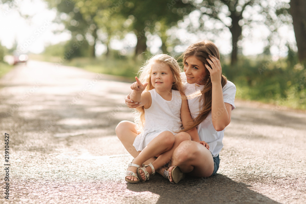 Attractive Mom and blonde hair daughter sits on road near big alley. They smile and look to natune Attractive Mom and blonde hair daughter sits on road near big alley. They smile and look to natune