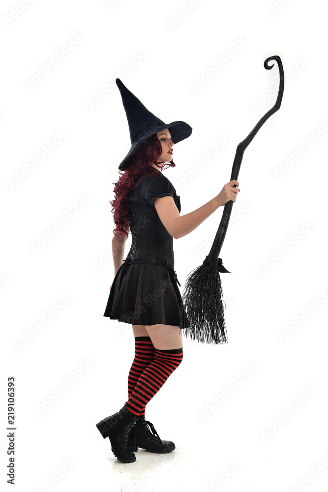 full length portrait of red haired girl wearing black witch costume and pointy hat, holding a broom. standing pose, isolated on white studio background. full length portrait of red haired girl wearing black witch costume and pointy hat, holding a broom. standing pose, isolated on white studio background.