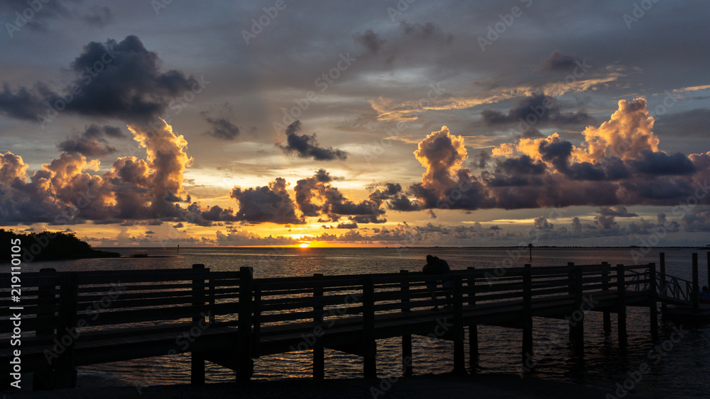 A couple in silhouette (unidentifiable) watches a beautiful Florida Gulf Coast sunset from a pier at Fort Island Beach, a popular spot from which to view these glorious sunsets.