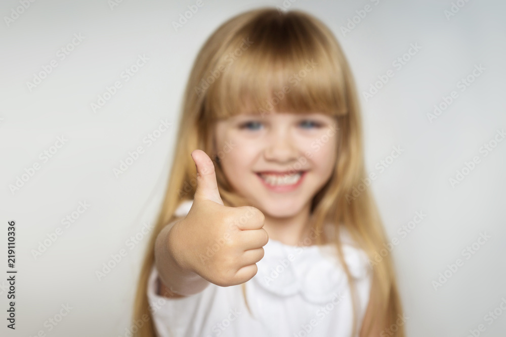 little blonde girl in white dress smiles and shows thumb up. focus on the finger little blonde girl in white dress smiles and shows thumb up. focus on the finger