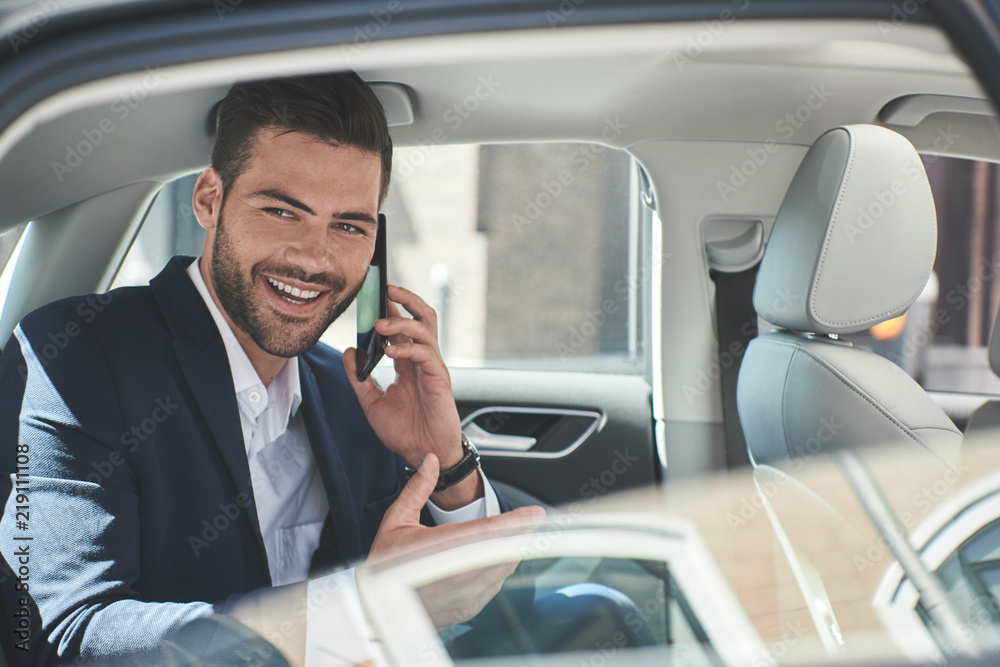 Young perfectionist. Handsome young man in full suit talking on smart phone while sitting in the car Young perfectionist. Handsome young man in full suit talking on smart phone while sitting in the car