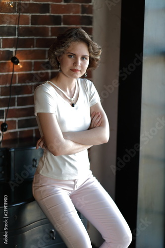 The brunette in light tight clothes posing against the background of an aged brickwork  hung with incandescent lamps.