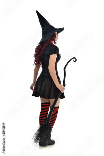 full length portrait of red haired girl wearing black witch costume and pointy hat, holding a broom. standing pose, isolated on white studio background.