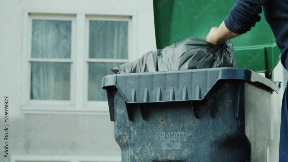 A man throws a bag of garbage in a garbage can near his house