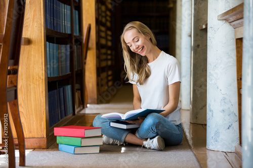 Young girl sitting on the floor in traditional old library at bookshelves, reading books.. Smiling and laughing student working, studying. Higher education. 
