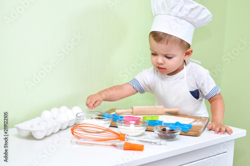 A cute two year old boy dressed as a chef makes cookies from Ingredients that are on his desk.