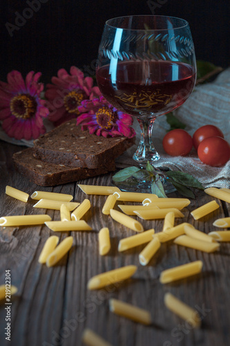 The pasta is scattered around the old wooden table. A glass of red wine is on the table. On the table pasta  tomatoes  parsley  basil  black bread  burlap  knife and fork.