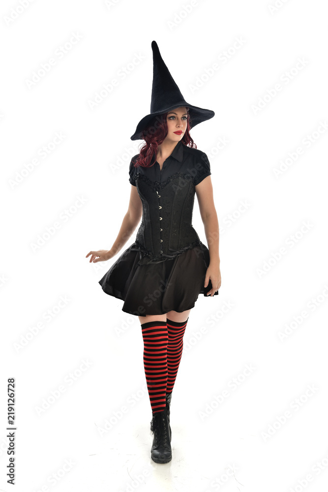 full length portrait of red haired girl wearing black witch costume and pointy hat.  standing pose, isolated on white studio background.