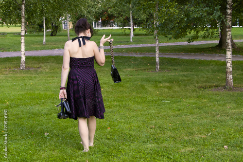 Portrait of a seductive young brunette in high heels smiling, walking barefoot on fresh grass in a sunny spring park, in a stylish black dress