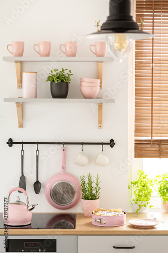 Real photo of kitchen interior with pastel pink mugs and bowls on shelves, fresh plants and homemade sponge cake on countertop