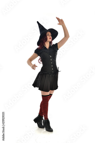 full length portrait of red haired girl wearing black witch costume and pointy hat. standing pose, isolated on white studio background.