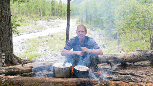A man is sitting near a fire with pots. The tourist cooks food at the stake, sits on a fallen tree.