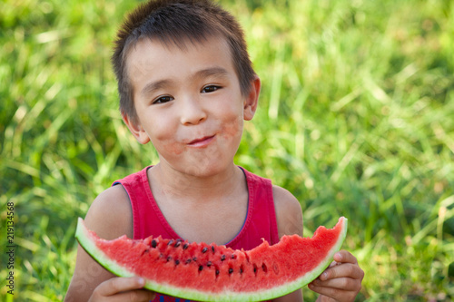 Happy asian child eating watermelon in the garden. Kids eat fruit outdoors. Healthy snack for children. Little boy playing in the garden biting a slice of watermelon.