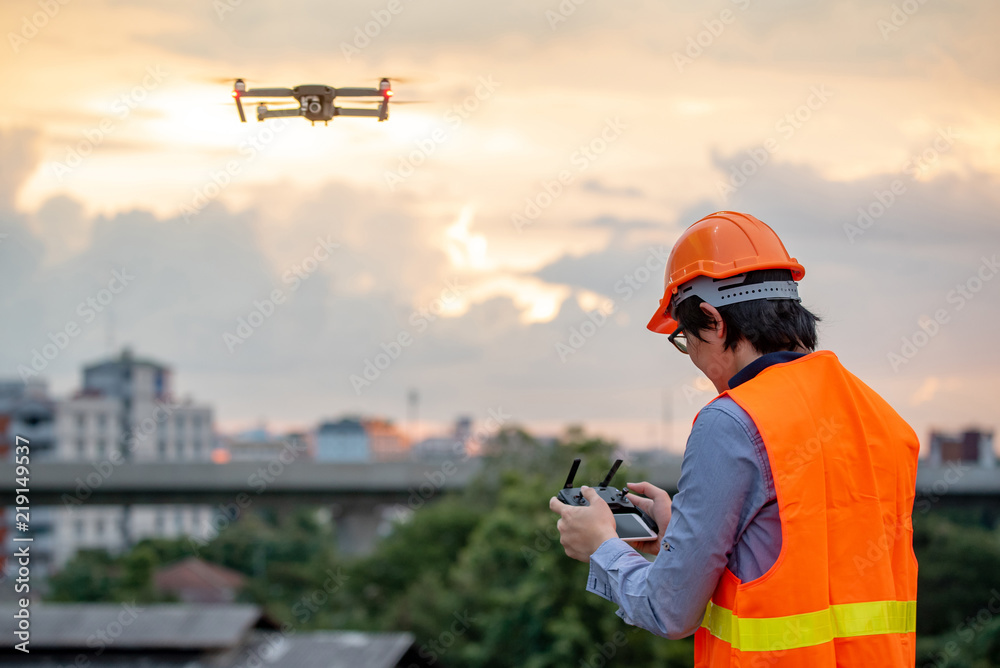 Young Asian engineer flying drone over construction site during sunset. Using unmanned aerial vehicle (UAV) for land and building site survey in civil engineering project. Young Asian engineer flying drone over construction site during sunset. Using unmanned aerial vehicle (UAV) for land and building site survey in civil engineering project.