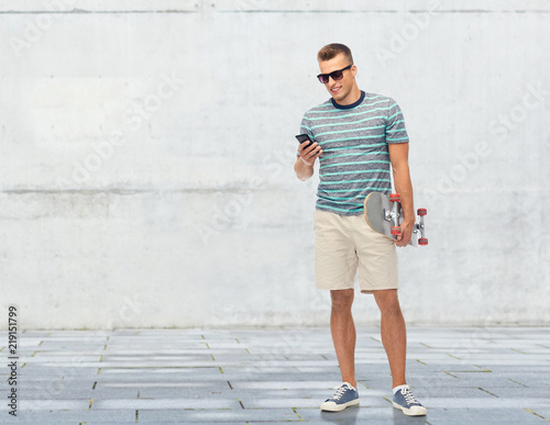Fototapeta Naklejka Na Ścianę i Meble -  technology, leisure and skateboarding concept - smiling young man with skateboard and smartphone over concrete wall background