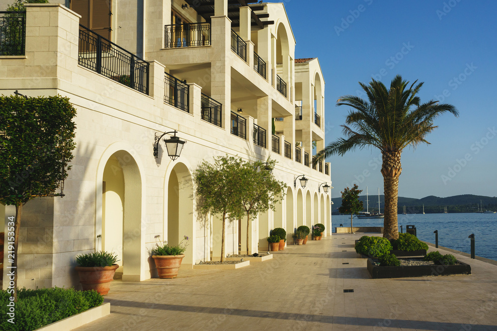 White stone embankment with palm trees, modern luxury architecture and large yachts in the background. 