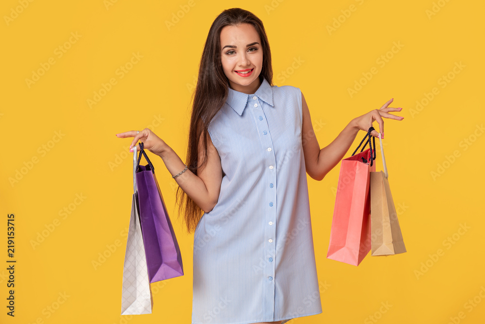 Image of a young brunette lady in white and blue summer striped dress posing with shopping bags and looking at camera over yellow background.