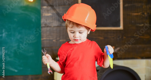 Toddler in protective hard hat, helmet at home in workshop. Child cute playing with wrench and toy saw, as builder or repairer, repairing or handcrafting. Kid boy play as handyman. Childhood concept.