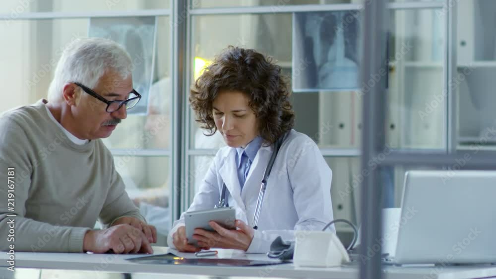 Beautiful female practitioner sitting at desk in modern doctor office with glass walls and discussing something on digital tablet with senior male patient