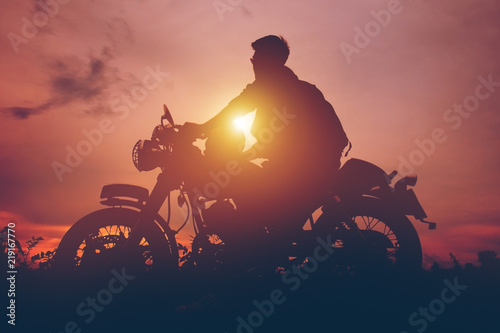 Silhouette of biker man  with his motorbike,he shoulder backpack. beside the natural lake and beautiful, enjoying freedom and active lifestyle, having fun on a bikers tour.sunset background and sky.