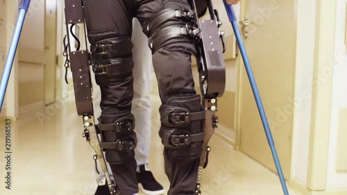 Legs of disable man in in the robotic exoskeleton walking through the corridor of the rehabilitation clinic. Doctor helping him. photo