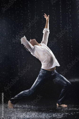 A young barefoot man wearing wet blue jeans and a white shirt expressively dances modern dances on the water under rainy waterdrops among the watersplashes
