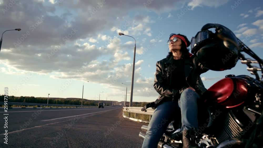 A biker old woman in a leather jacket and gloves is sitting on her cool motorcycle. A woman in round glasses and a red bandana. There's an empty highway in the background. The life of an elderly A biker old woman in a leather jacket and gloves is sitting on her cool motorcycle. A woman in round glasses and a red bandana. There's an empty highway in the background. The life of an elderly