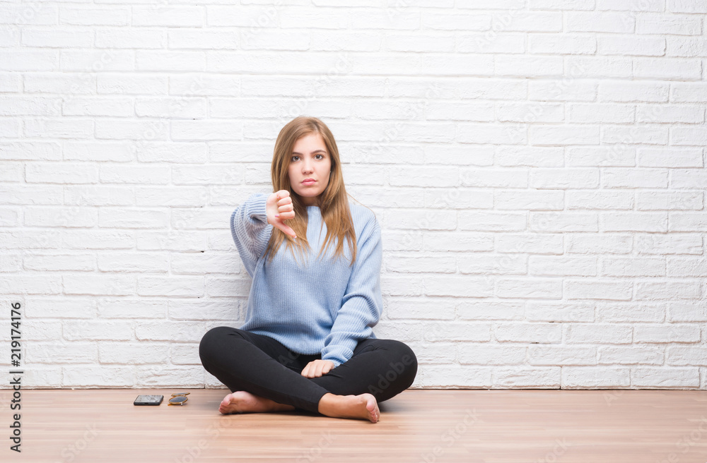 Young adult woman sitting on the floor in autumn over white brick wall looking unhappy and angry showing rejection and negative with thumbs down gesture. Bad expression. Young adult woman sitting on the floor in autumn over white brick wall looking unhappy and angry showing rejection and negative with thumbs down gesture. Bad expression.