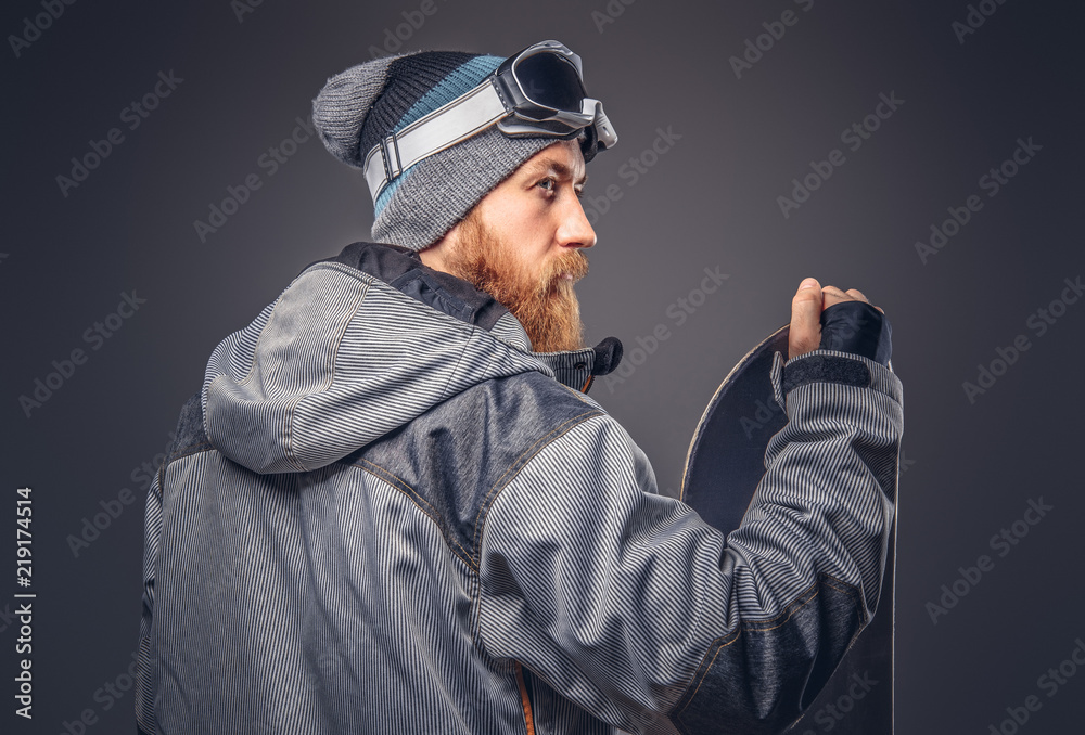 Brutal redhead snowboarder with a full beard in a winter hat and protective glasses dressed in a snowboarding coat posing with snowboard at a studio, looking away. Isolated on the gray background. Brutal redhead snowboarder with a full beard in a winter hat and protective glasses dressed in a snowboarding coat posing with snowboard at a studio, looking away. Isolated on the gray background.