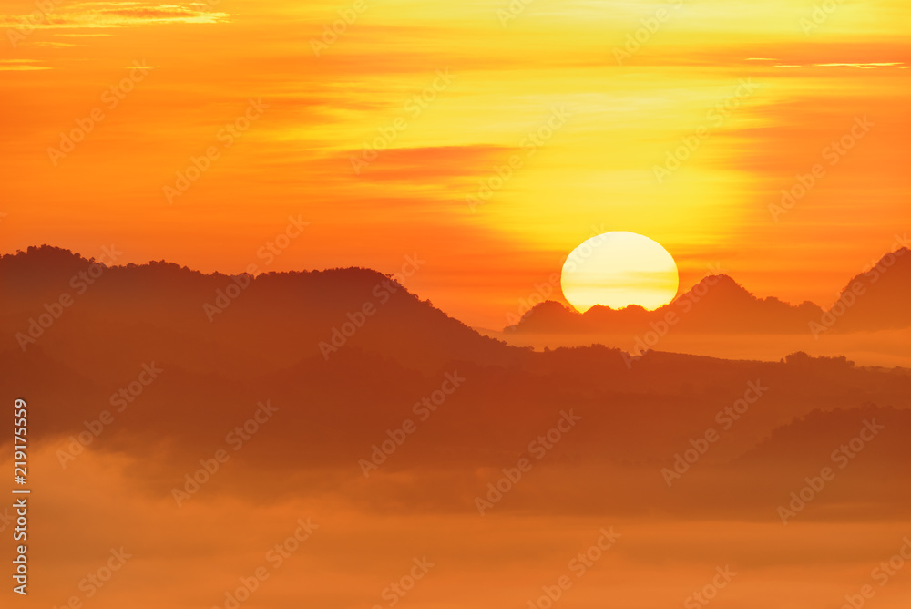 Distant scenic view at dawn / scene of evanescent atmosphere in the evergreen forest. Big sun rises behind a mountain range in the morning. Dense morning fog or mist rolling in over lush wilderness.
