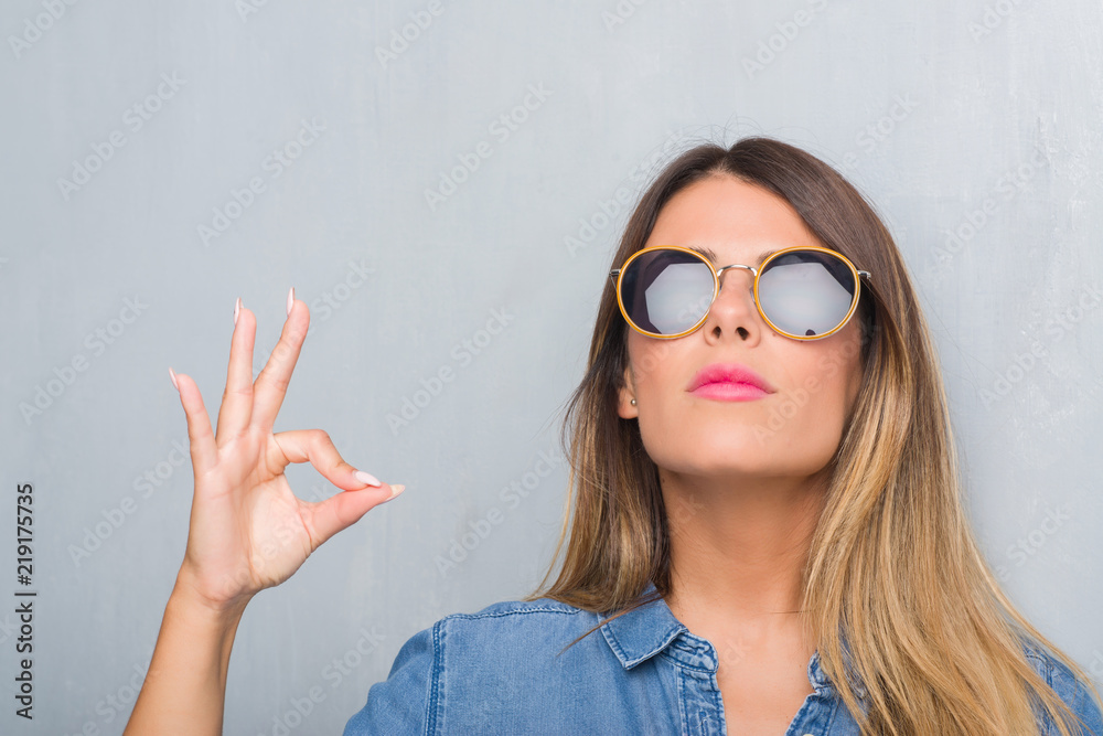 Young adult woman over grunge grey wall wearing retro sunglasses relax and smiling with eyes closed doing meditation gesture with fingers. Yoga concept. Young adult woman over grunge grey wall wearing retro sunglasses relax and smiling with eyes closed doing meditation gesture with fingers. Yoga concept.