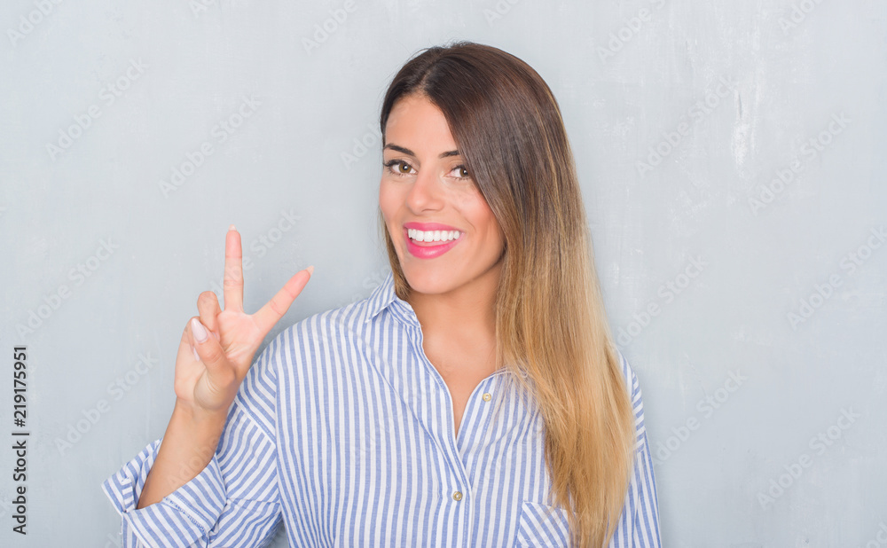 Young adult woman over grunge grey wall wearing business shirt smiling with happy face winking at the camera doing victory sign. Number two. Young adult woman over grunge grey wall wearing business shirt smiling with happy face winking at the camera doing victory sign. Number two.