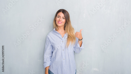 Young adult woman over grey grunge wall wearing fashion business outfit smiling with happy face looking and pointing to the side with thumb up. © Krakenimages.com