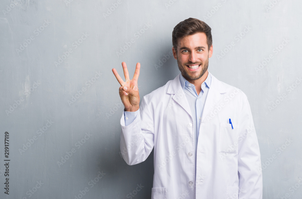 Handsome young professional man over grey grunge wall wearing white coat showing and pointing up with fingers number three while smiling confident and happy. Handsome young professional man over grey grunge wall wearing white coat showing and pointing up with fingers number three while smiling confident and happy.